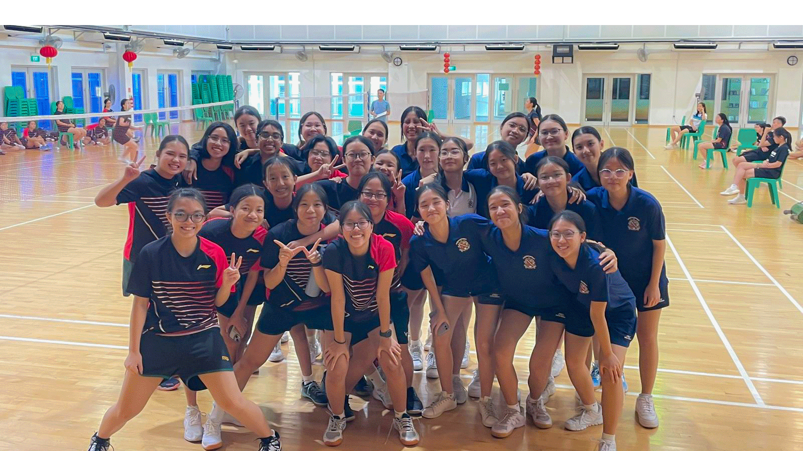 A large group of young female students in sports uniforms posing together on an indoor badminton court.