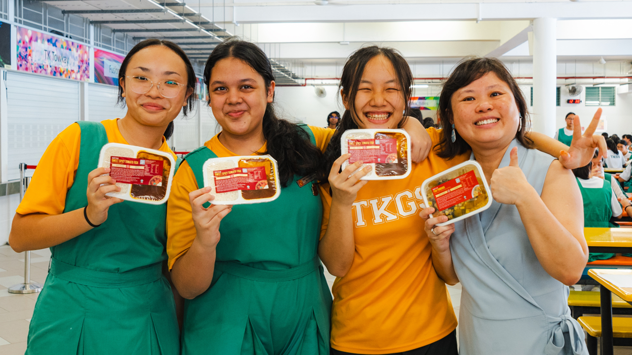 Four smiling people in a school canteen holding packaged food trays, three wearing TKGS uniforms.