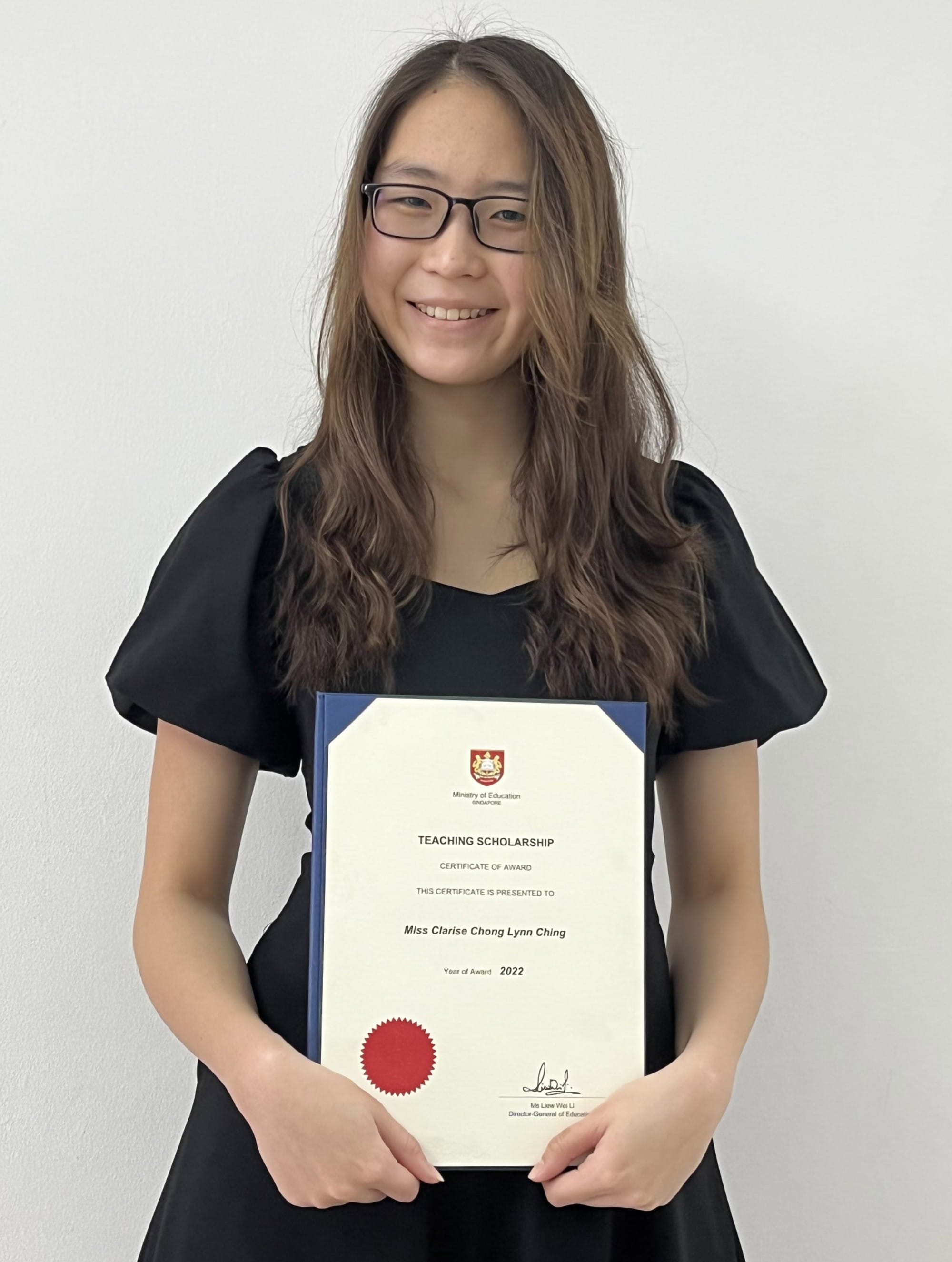 A smiling young woman with glasses holds a 2022 Ministry of Education Teaching Scholarship certificate.