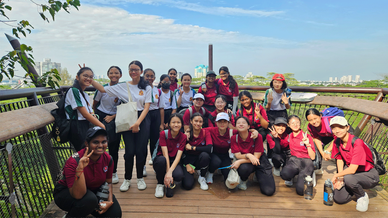 A large group of young students in red and white uniforms pose on an outdoor wooden deck with a city skyline behind them.