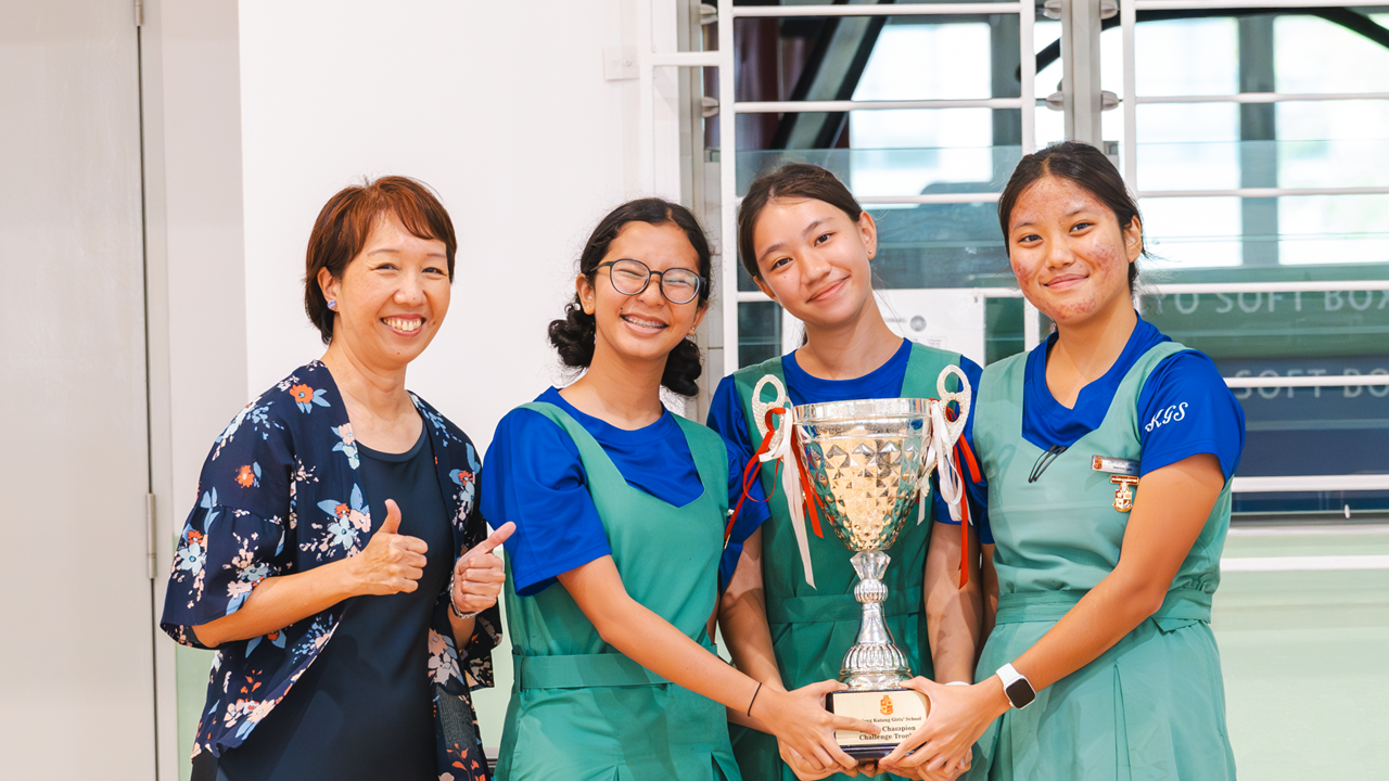 Four smiling females, one adult and three students in blue and green uniforms, holding a trophy with ribbons.