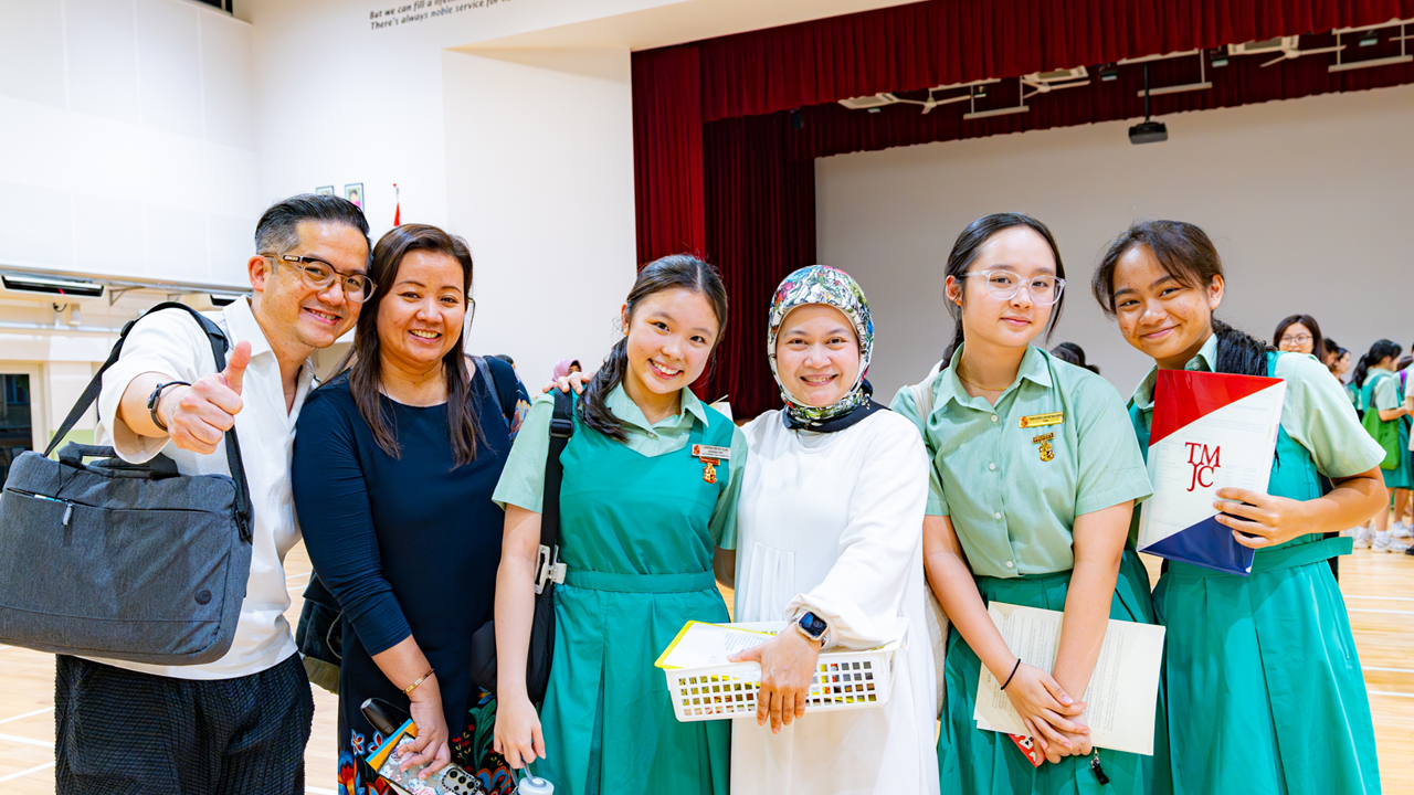 Six people smile together in a school hall, four girls in green uniforms stand with two adults, one giving a thumbs up.