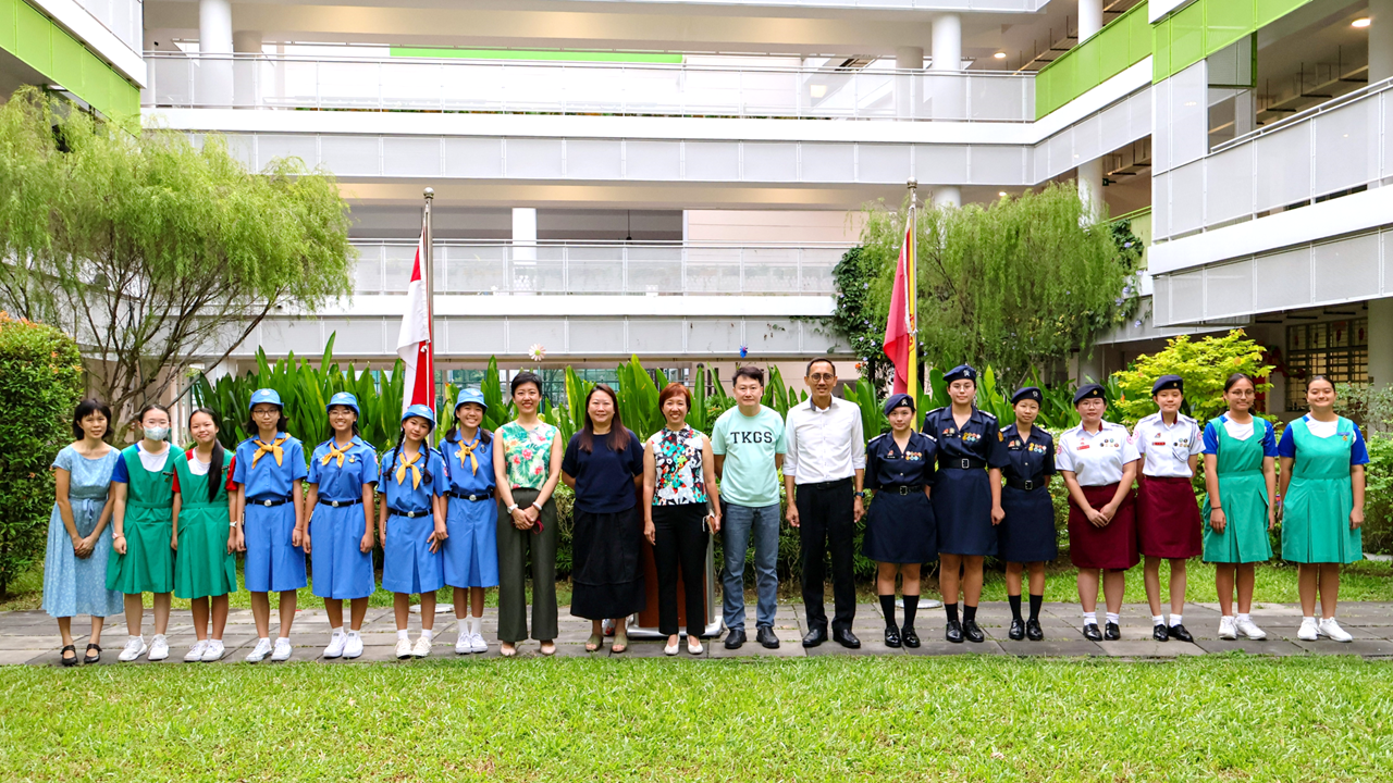 A group of students in various uniforms and adults pose outdoors in front of a multi-story school building.