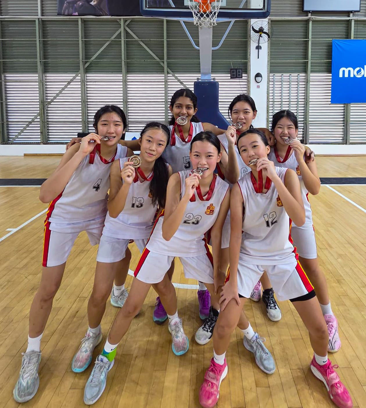 Eight young female basketball players in white uniforms pose together on a court, each biting their medals.