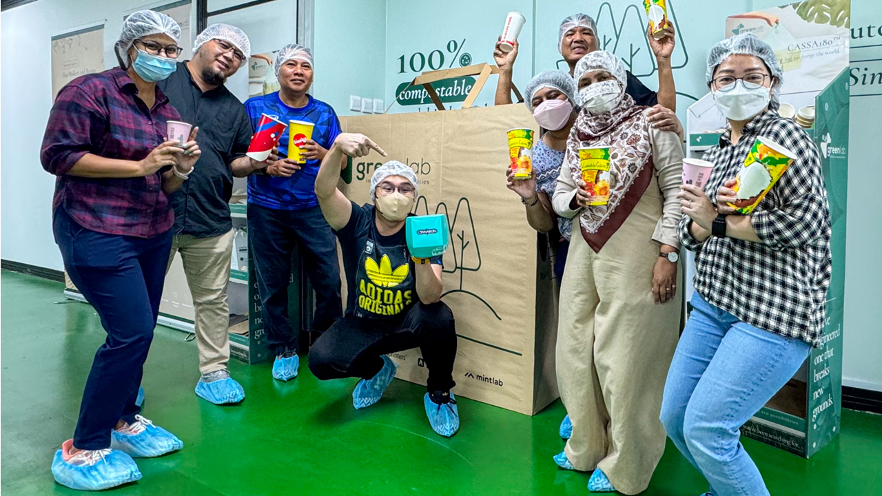 Group of people wearing hair nets and masks holding colorful cups near a large Greenlab compostable packaging box.