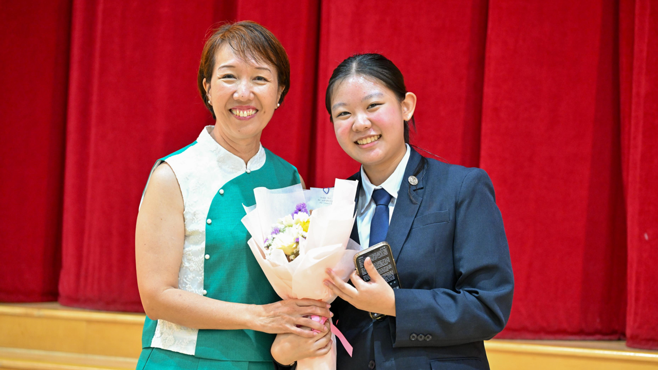 Two smiling women pose together holding a flower bouquet in front of a red curtain on a stage.