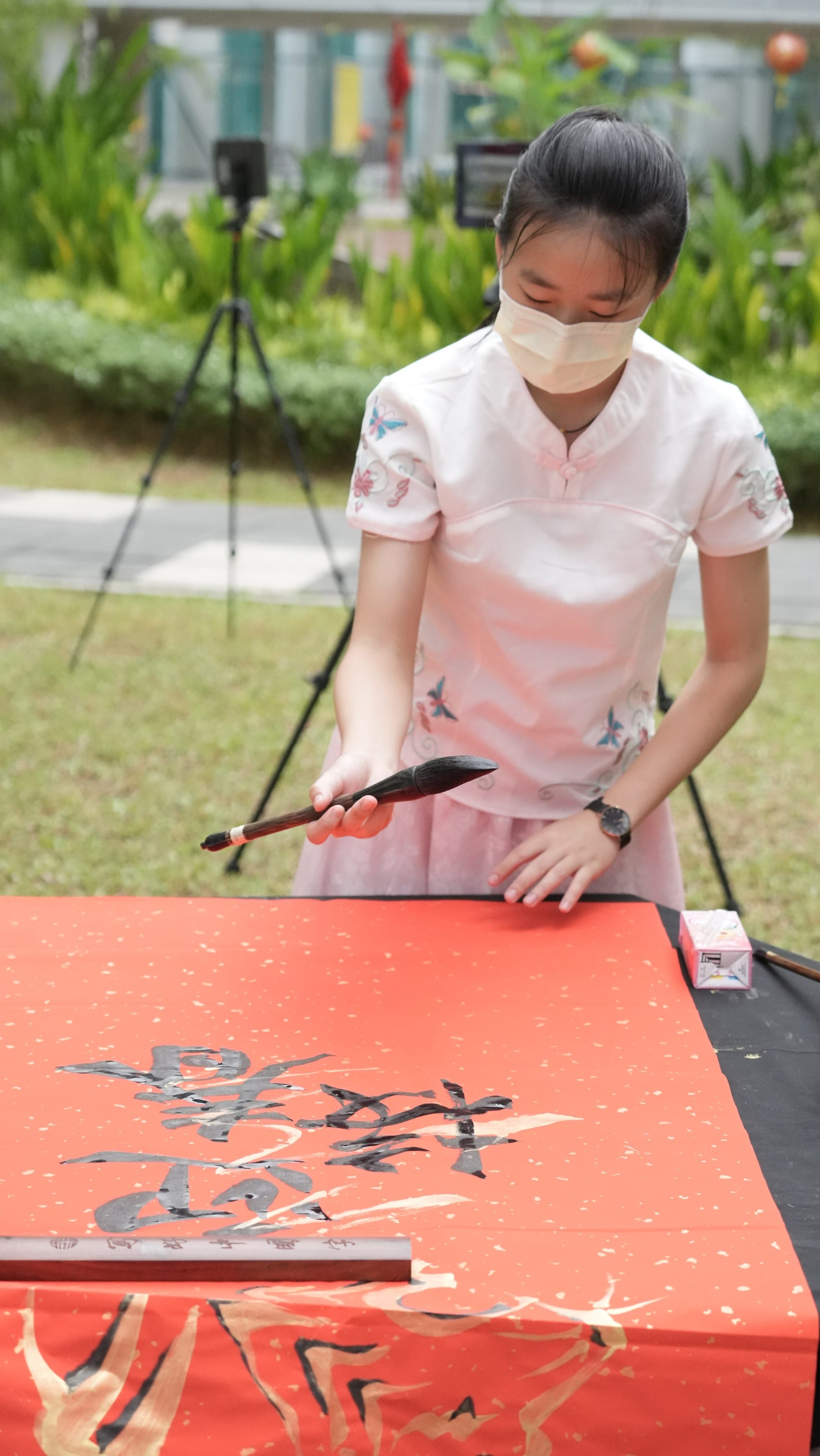 A young woman wearing a mask writes Chinese calligraphy with a brush on a red paper outdoors.