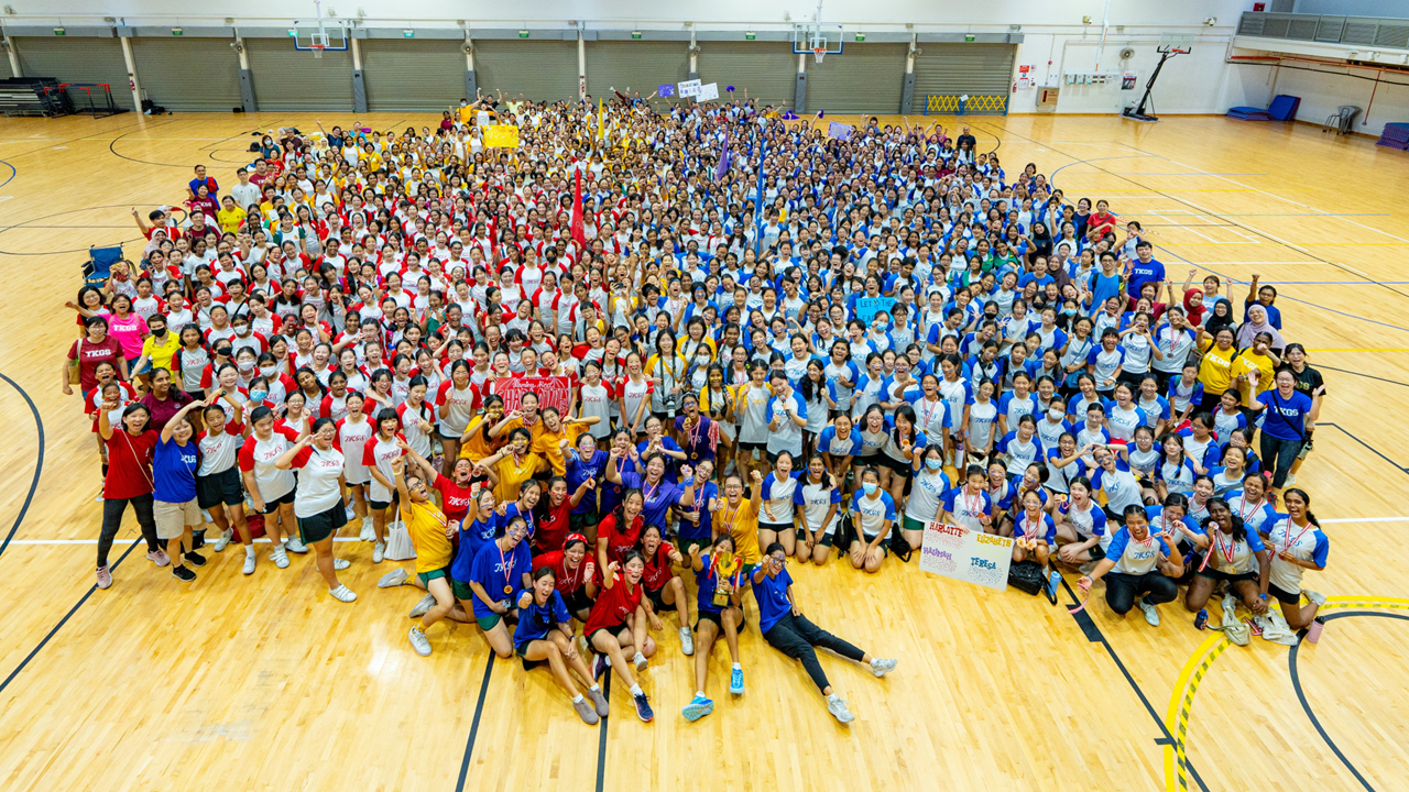 Large group of students in red, blue, yellow, and white shirts posing together on an indoor basketball court.