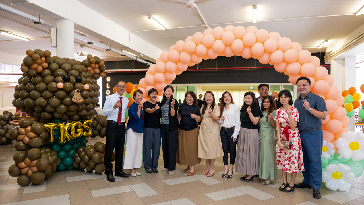A group of people posing under a peach balloon arch next to a large bear balloon sculpture labeled TKGS.