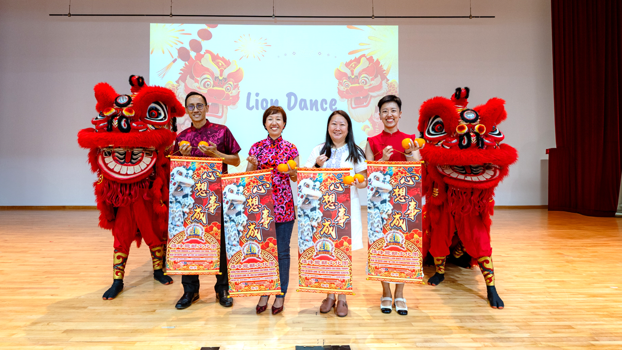 Four people holding banners and oranges pose between two red lion dance costumes on a stage with a Lion Dance backdrop.