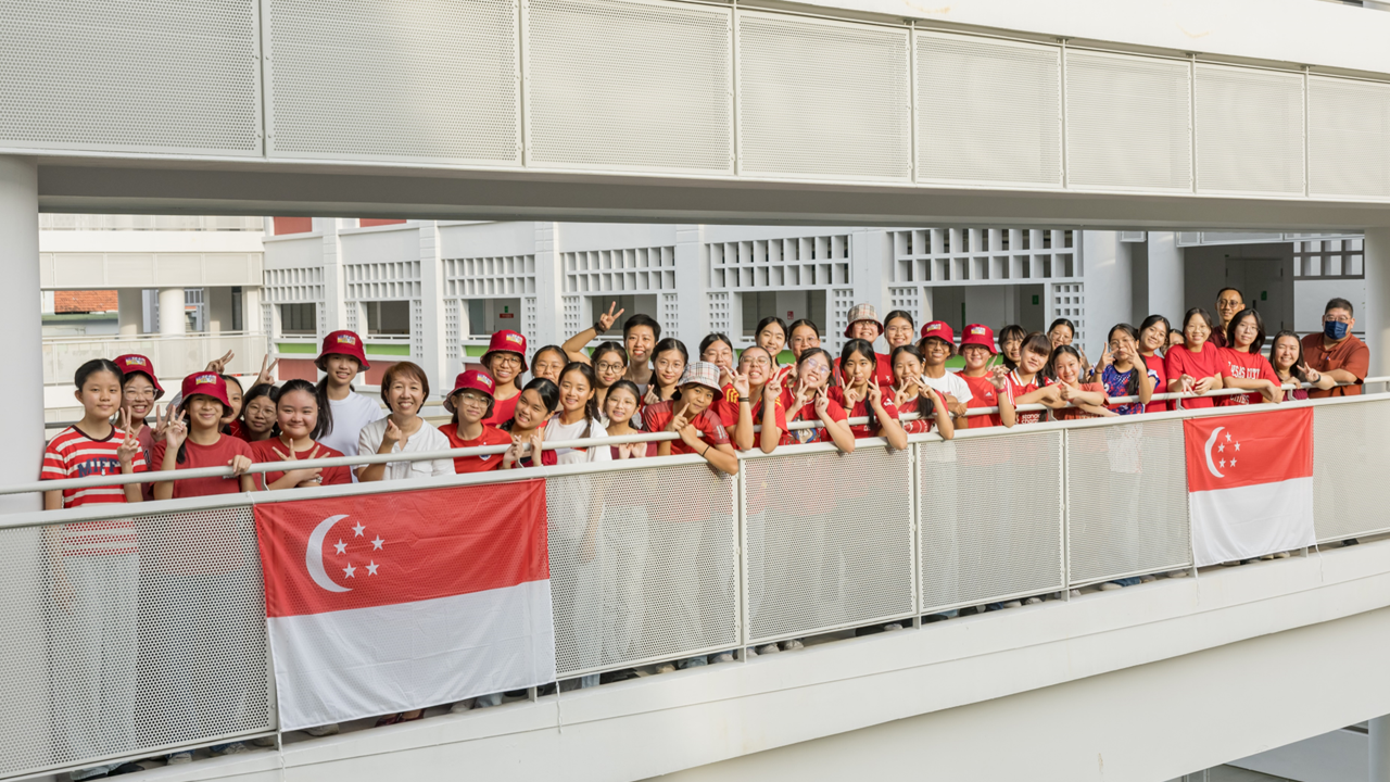 A large group of children and adults in red clothing pose on a balcony with two Singapore flags displayed.