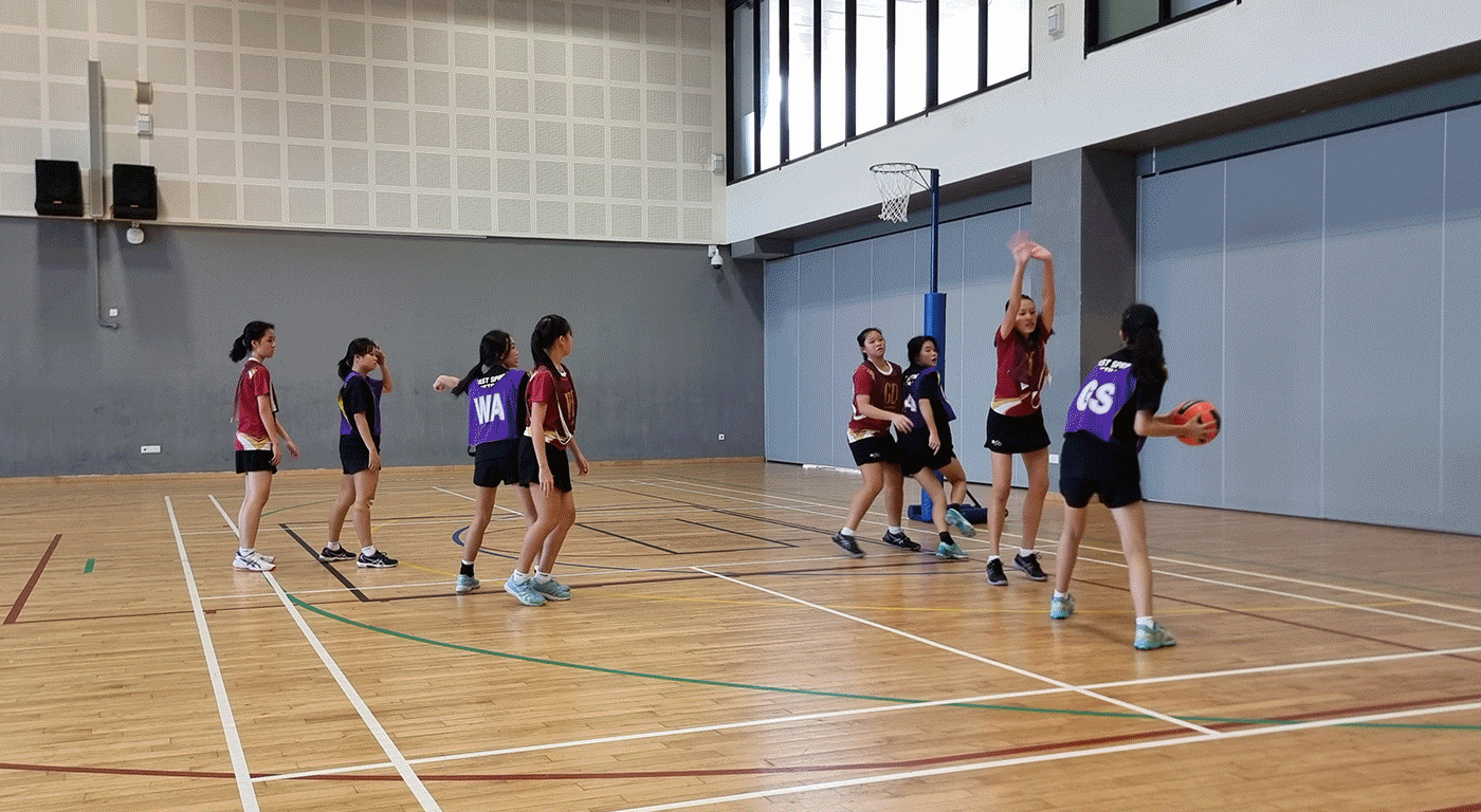 Girls in red and purple bibs playing netball indoors, one player jumping with arms raised near the hoop.