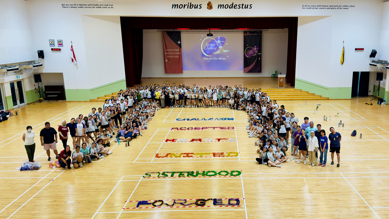Large group of students in a school hall surround colorful words on the floor spelling CHALLENGED, ACCEPTED, HEART, UNITED, SISTERHOOD, FORGED.