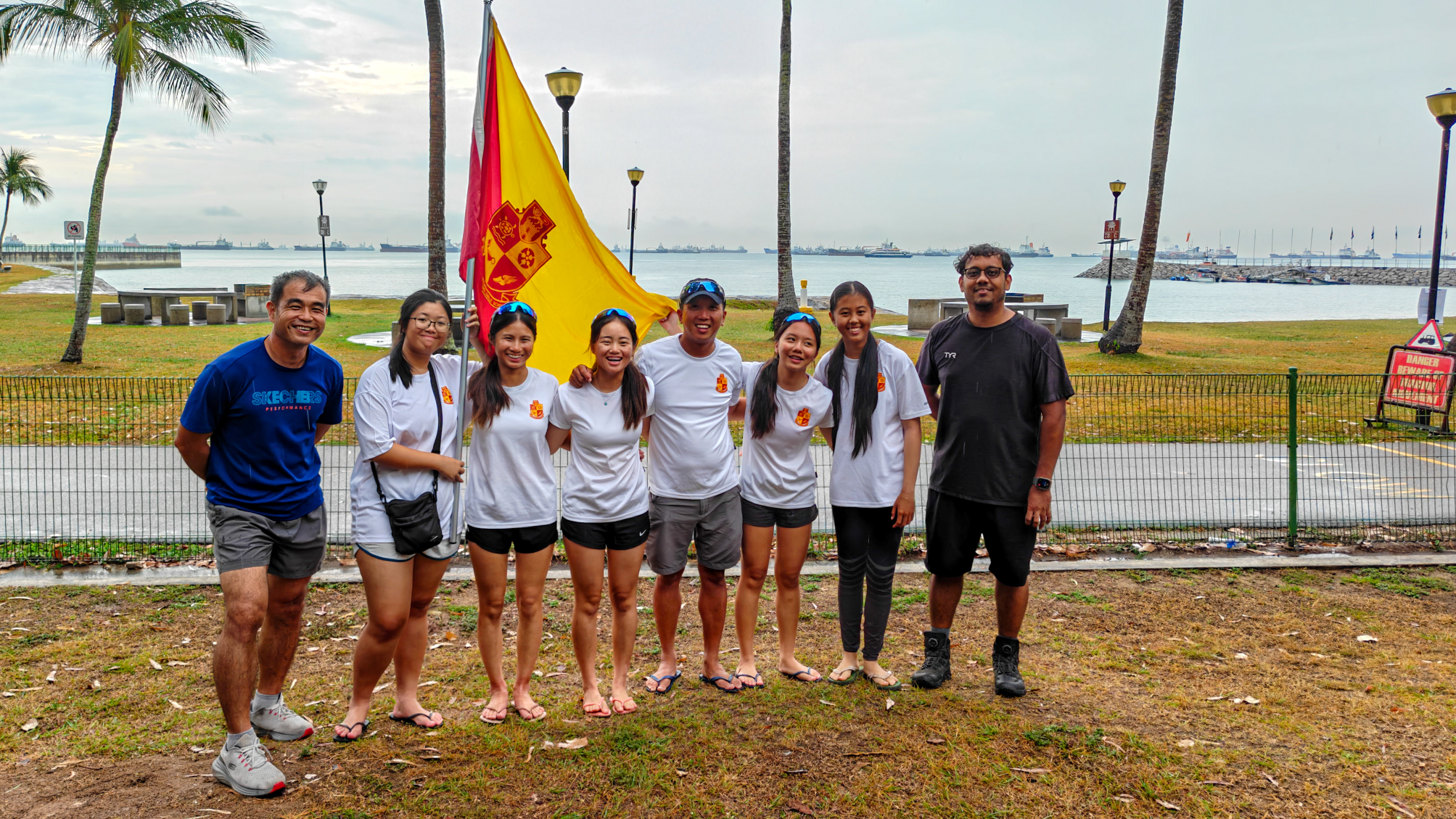 Eight people pose by the waterfront with palm trees, holding a yellow and red flag.