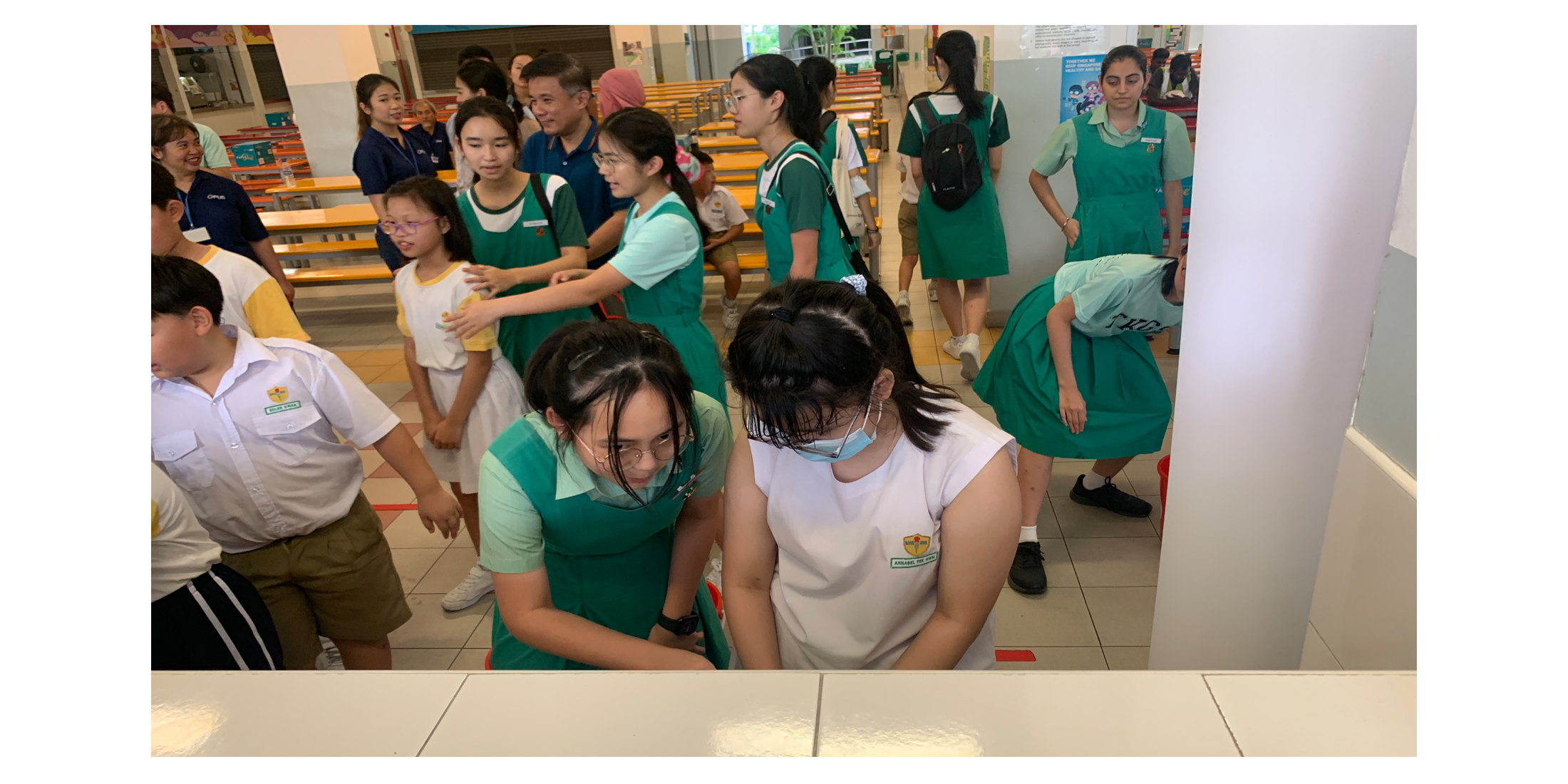 Students in green uniforms gather in a school canteen, with two girls crouching together in the foreground.