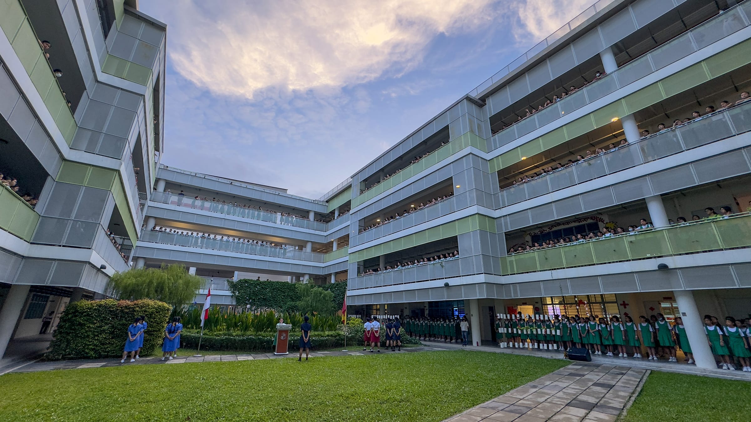Students in uniforms stand in a school courtyard during an assembly, with crowded multi-story balconies overlooking them.