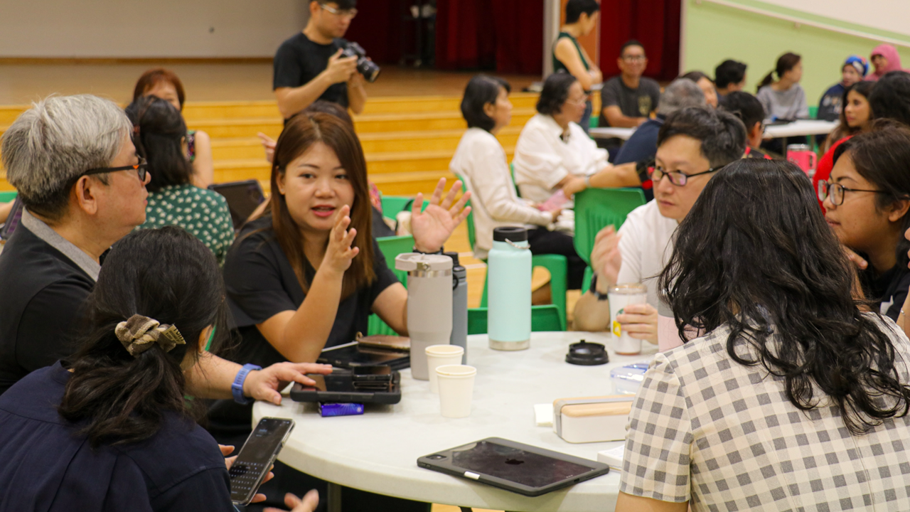 A group of people sit around a round table in a hall, talking, with cups and a tablet on the table.