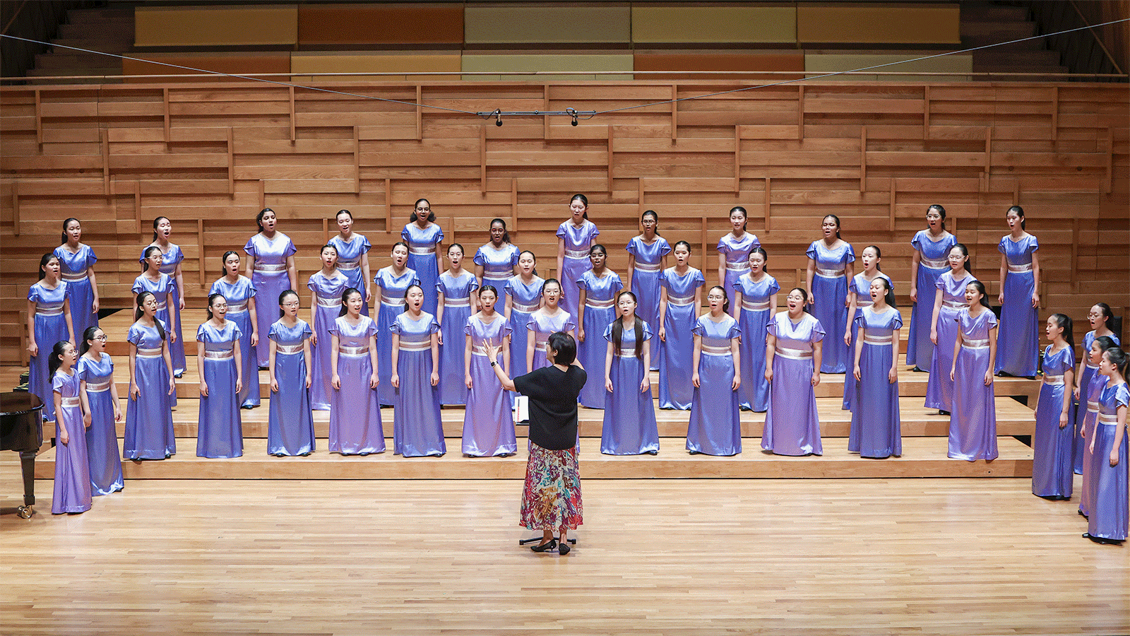 A conductor leads a large girls choir in purple satin dresses on a wooden concert hall stage.