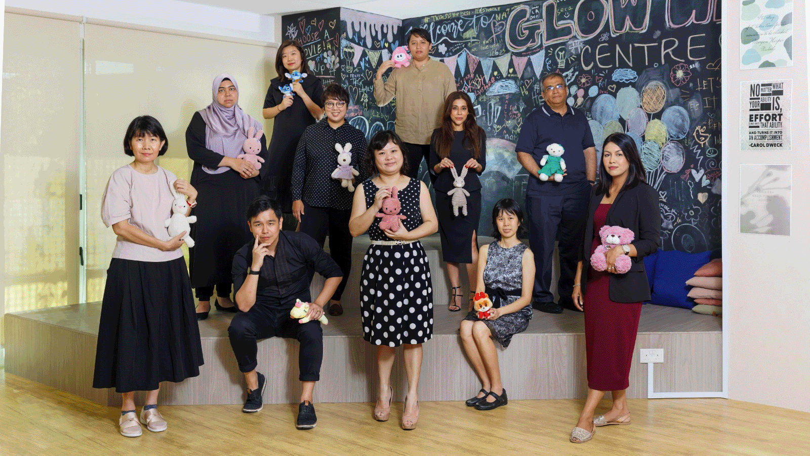 Group of 10 people posing indoors, each holding a stuffed toy, in front of a colorful chalkboard wall.