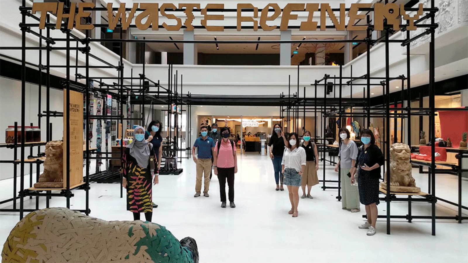 People wearing face masks stand inside The Waste Refinery exhibition with black scaffolding and cardboard sculptures.
