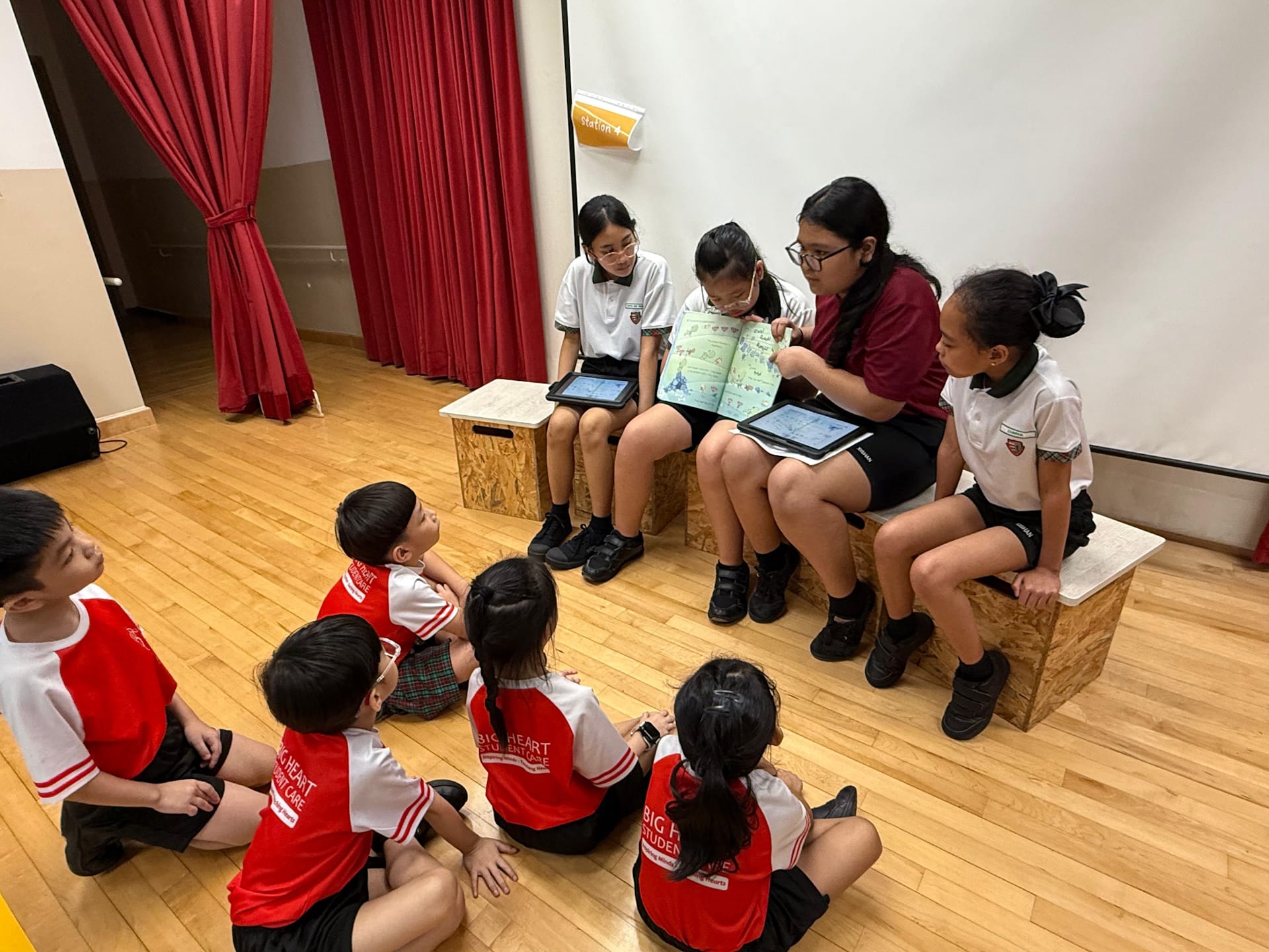 Older students sit on wooden benches reading a colorful book to young children seated on the floor in red uniforms.