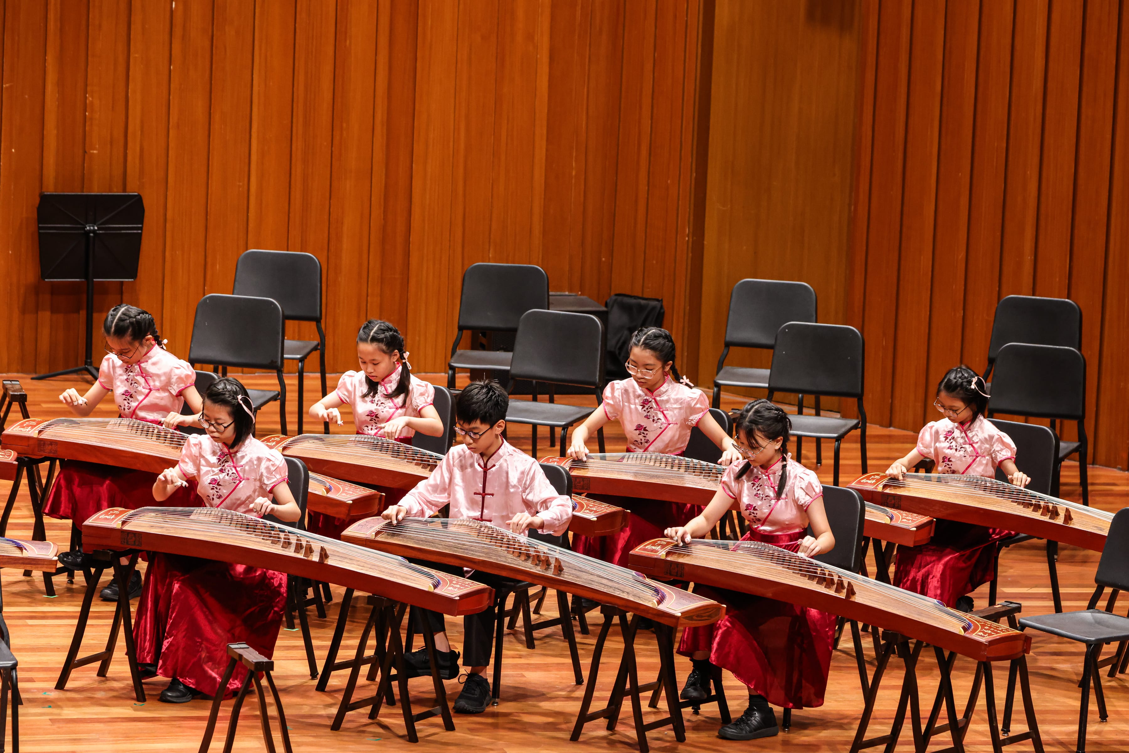 Children in pink and red traditional attire perform on guzheng instruments on a wooden stage.