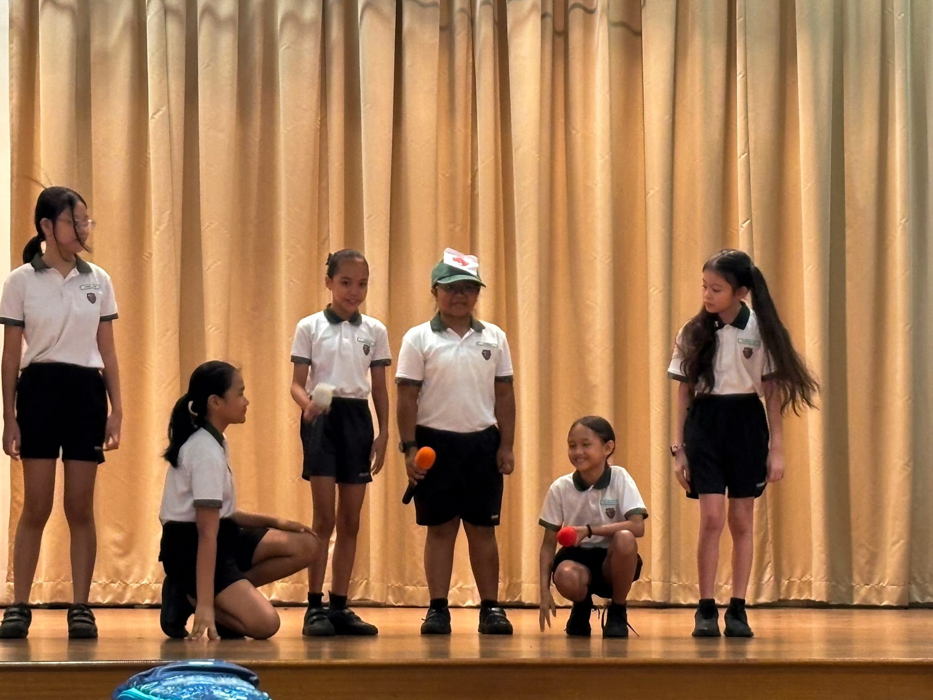 Six children in white and black school uniforms perform on a wooden stage in front of a beige curtain.