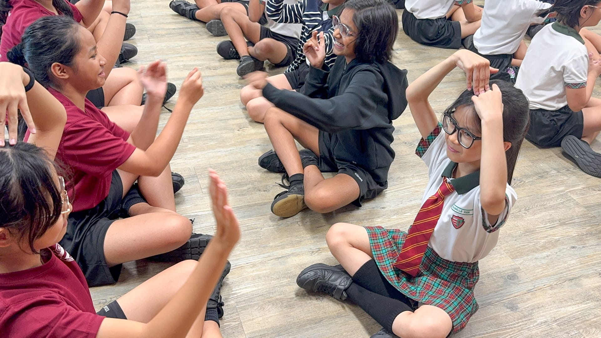 Children in school uniforms sit on a wooden floor, raising their hands and gesturing during a group activity.