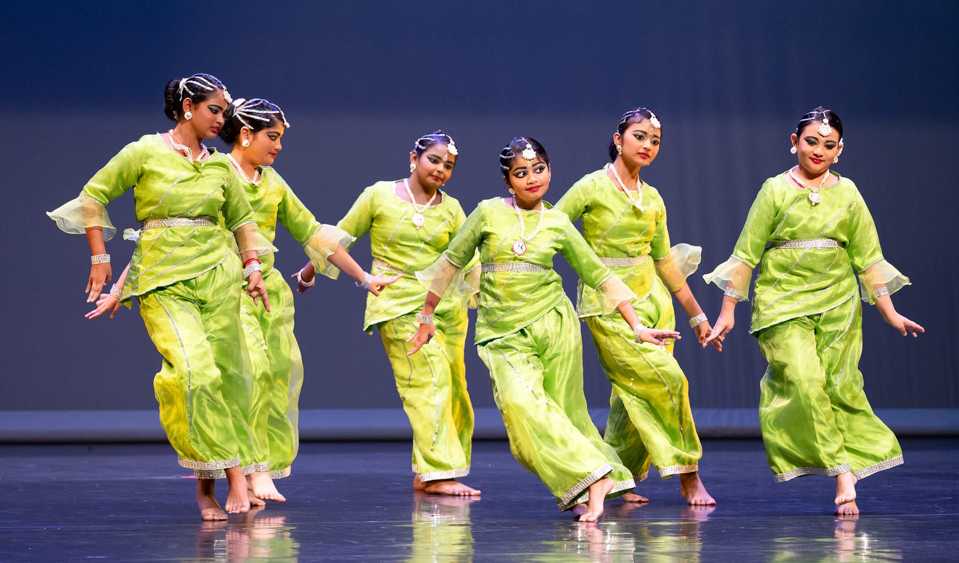 Six young women in matching lime green traditional Indian outfits perform a classical dance on stage.