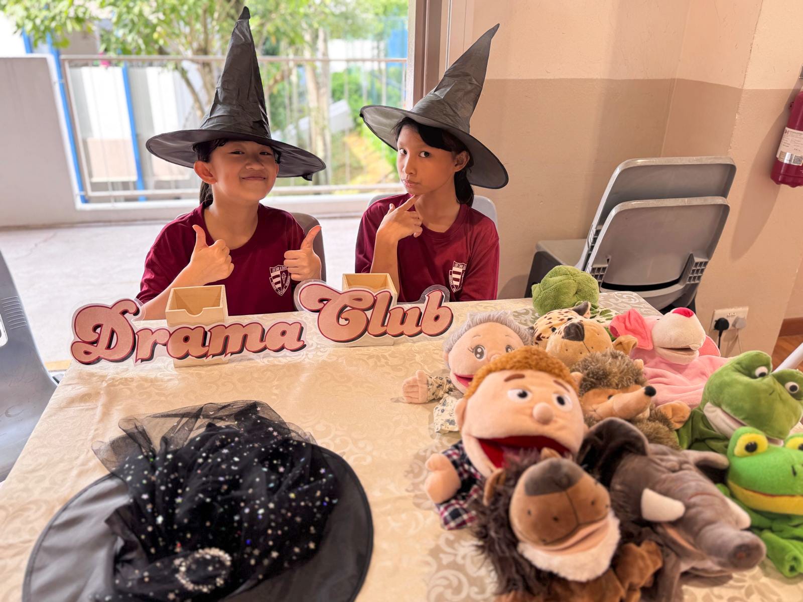 Two children wearing witch hats sit behind a Drama Club sign with hand puppets on the table.