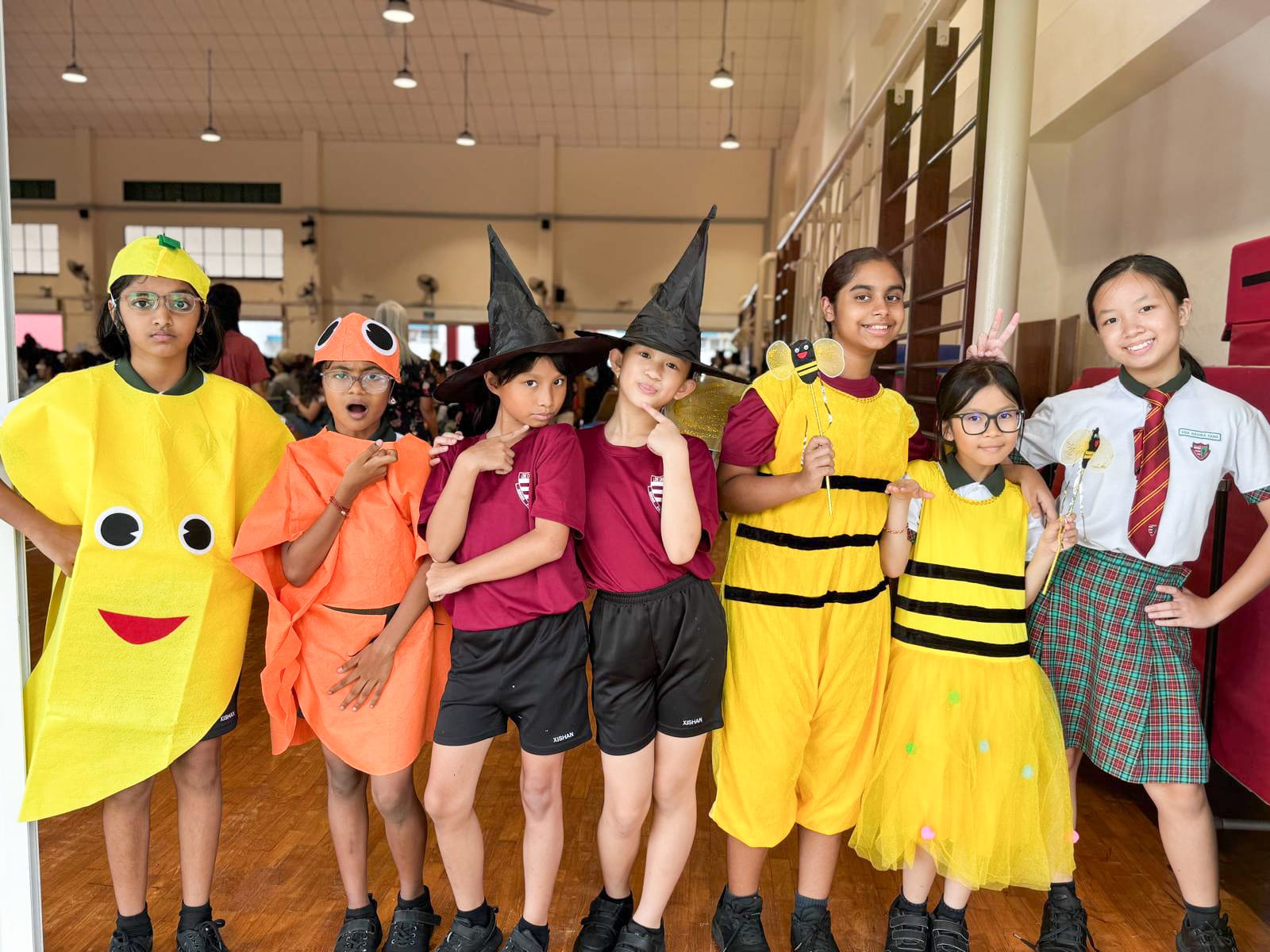 Seven children pose in costumes including a banana, fish, witches, and bees inside a school hall.
