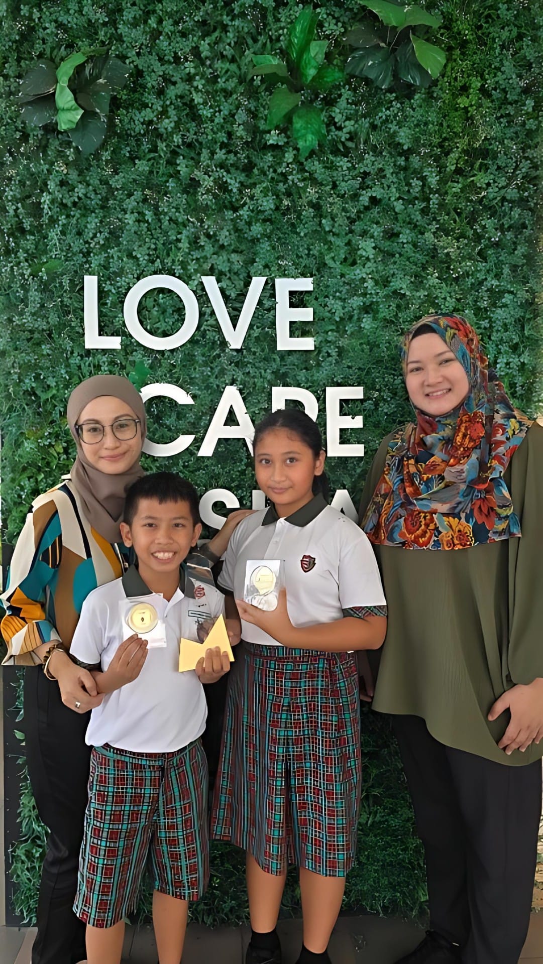 Two women wearing hijabs and two children in school uniforms pose in front of a green wall reading "Love Care."