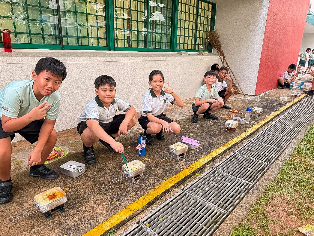 Four uniformed school boys crouch outdoors near small cooking containers on the ground, smiling at the camera.