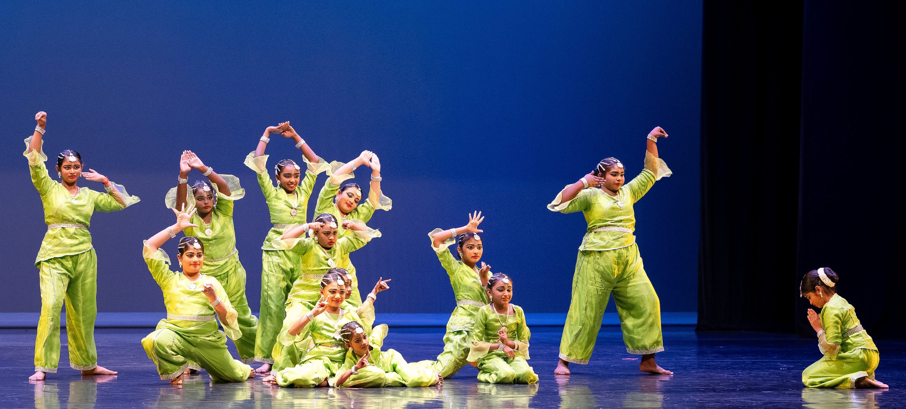 A group of dancers in bright green costumes perform a classical Indian dance on a blue-lit stage.