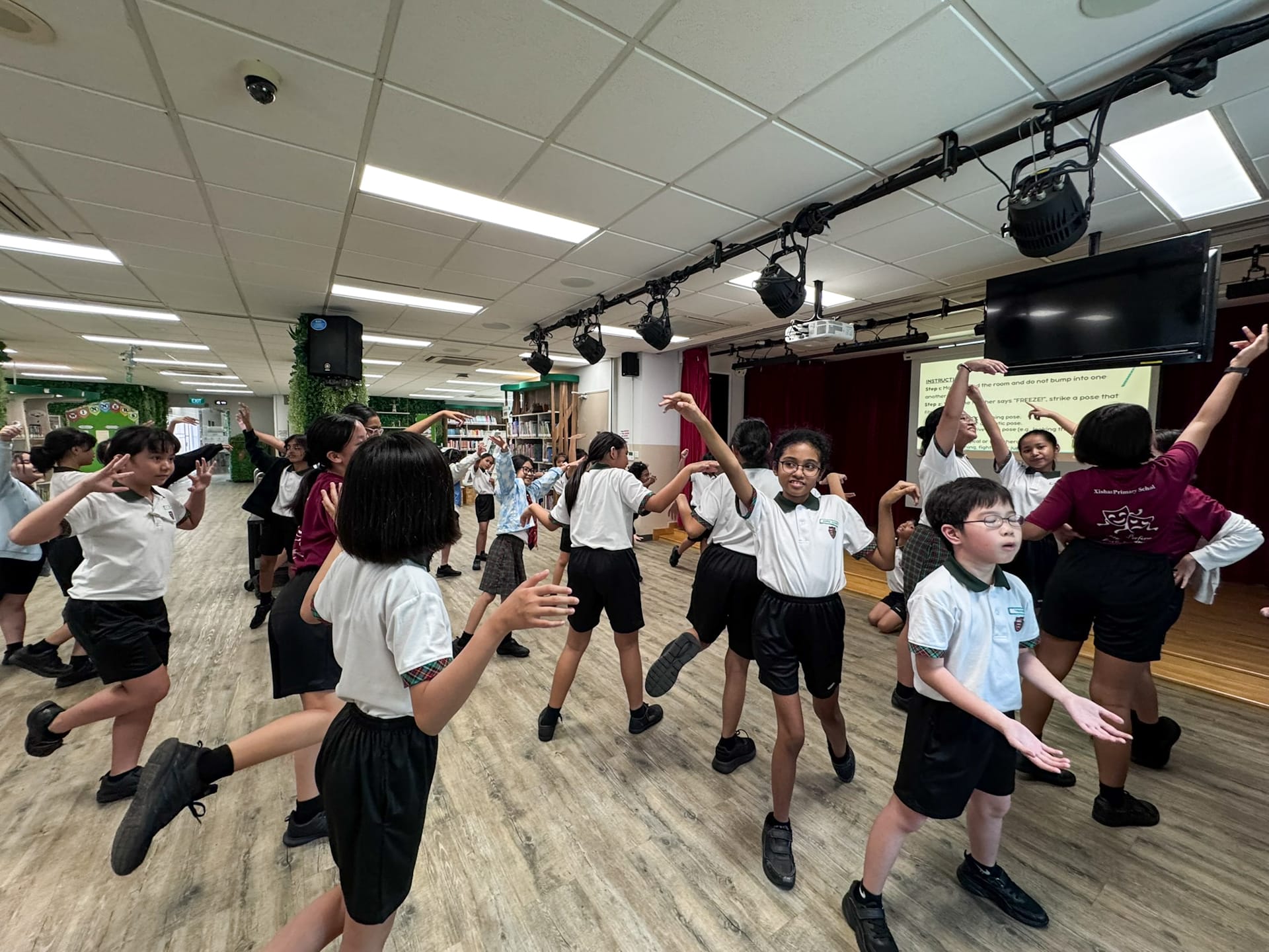 A group of primary school children in uniforms dancing and posing in a school hall with stage lights overhead.