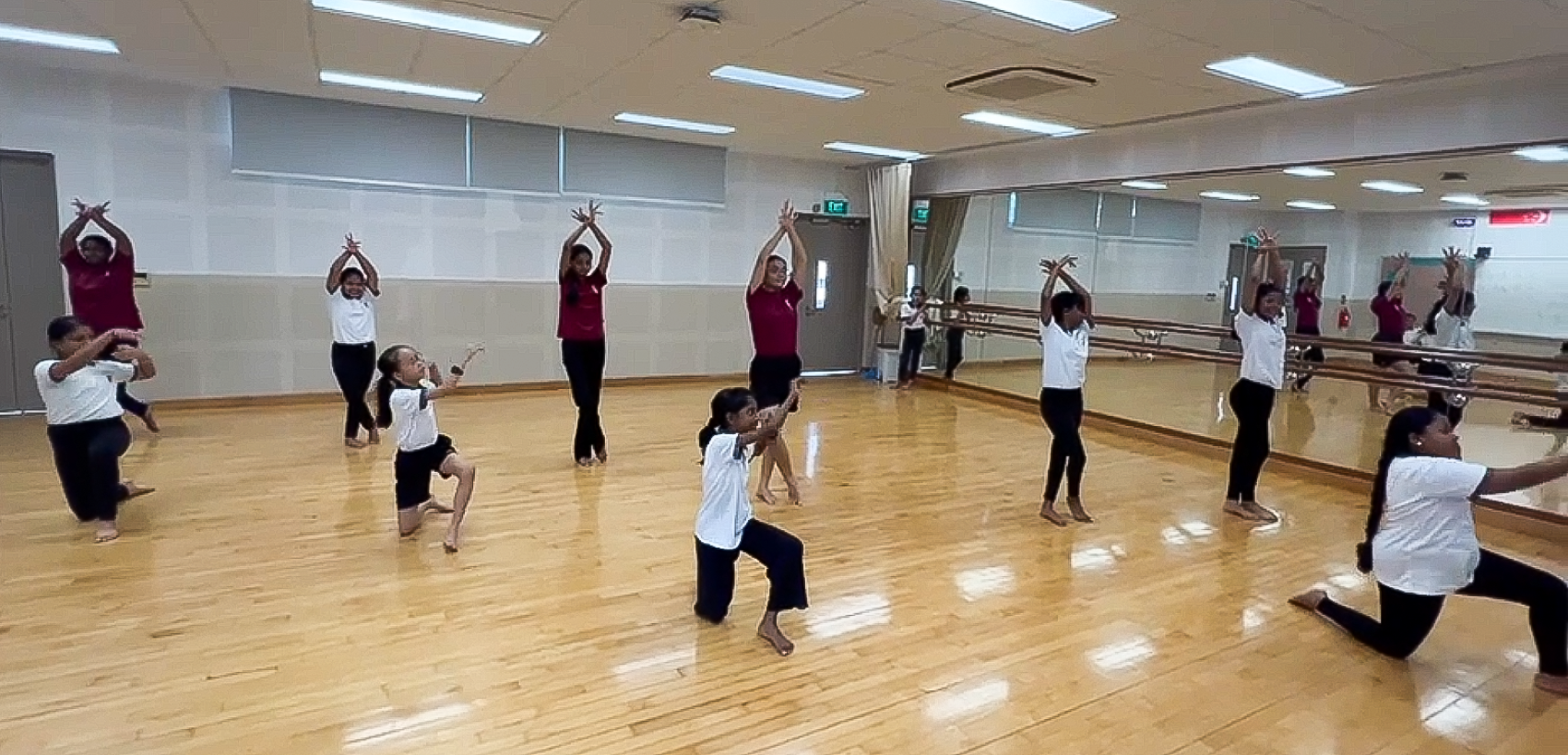 Children in white and maroon uniforms practice a dance routine in a mirrored studio with wooden floors.