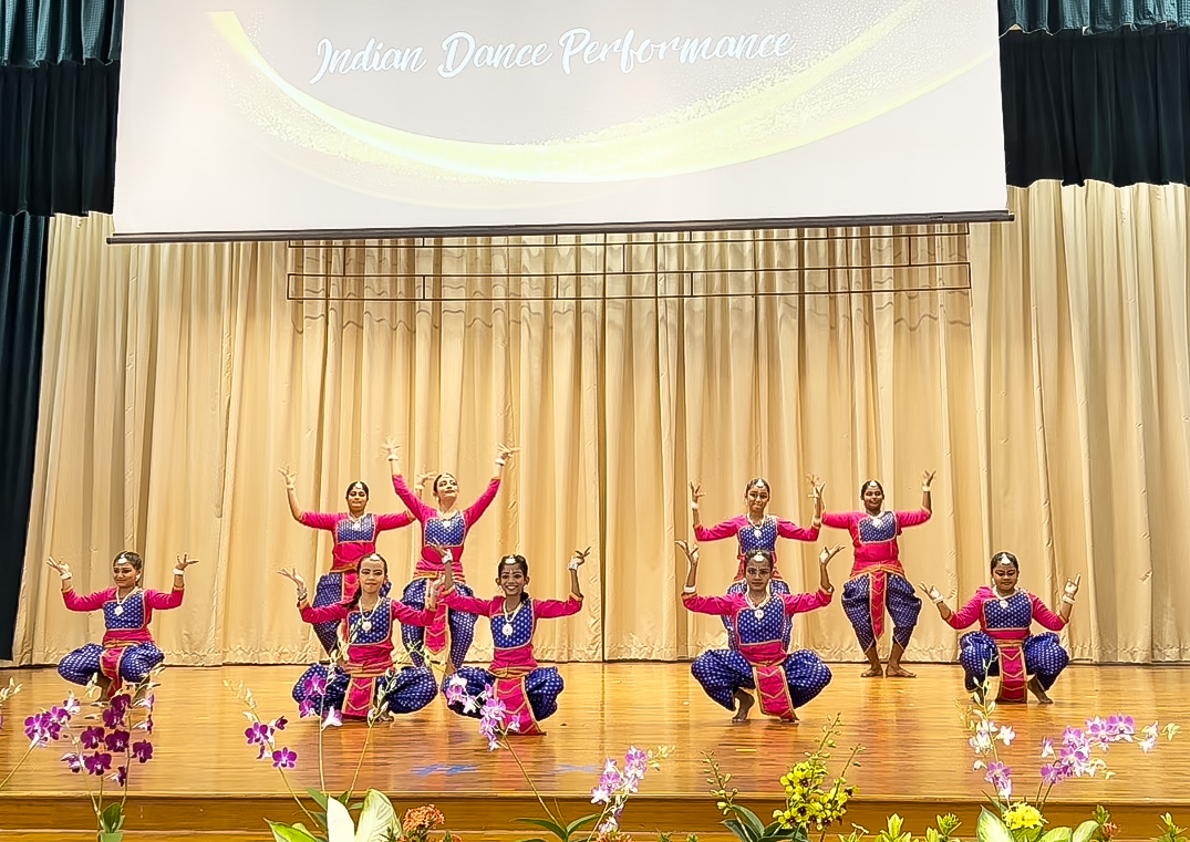 Nine dancers in pink and blue costumes perform on stage under a screen reading Indian Dance Performance.