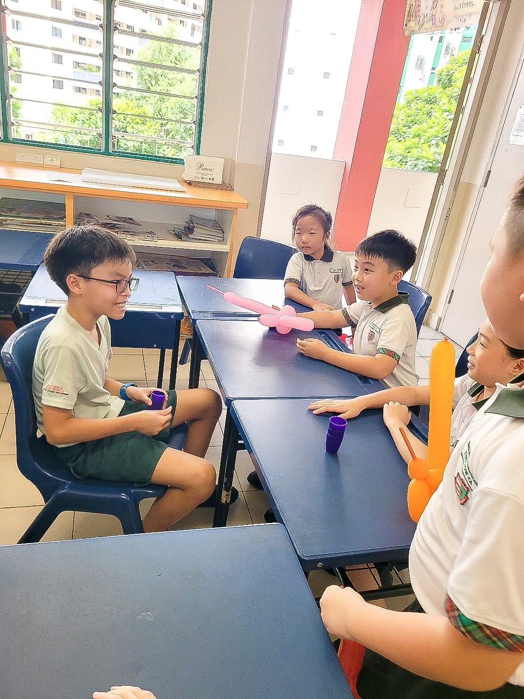 Four children in school uniforms sit around blue desks with twisted balloon animals and small cups on the table.