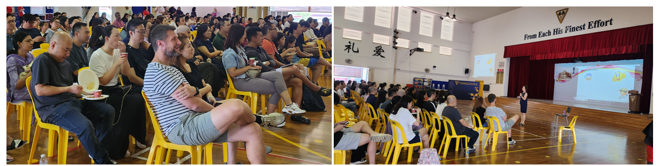 A crowd sits on yellow chairs in a school hall while a presenter speaks on stage before a projection screen.