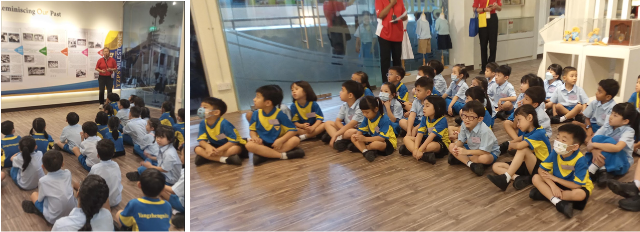 Children in blue and yellow uniforms sit on a wooden floor listening to a presenter in a museum exhibit hall.