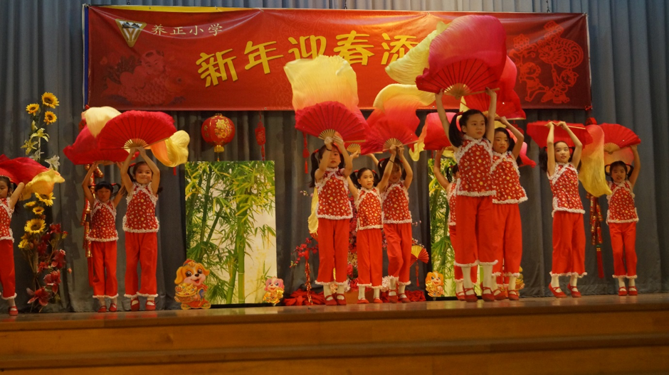 Children in red outfits perform a fan dance on stage under a Chinese New Year banner.