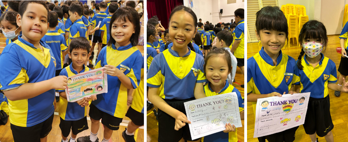 Six children in blue and yellow school uniforms smile while holding handmade Thank You cards in a school hall.