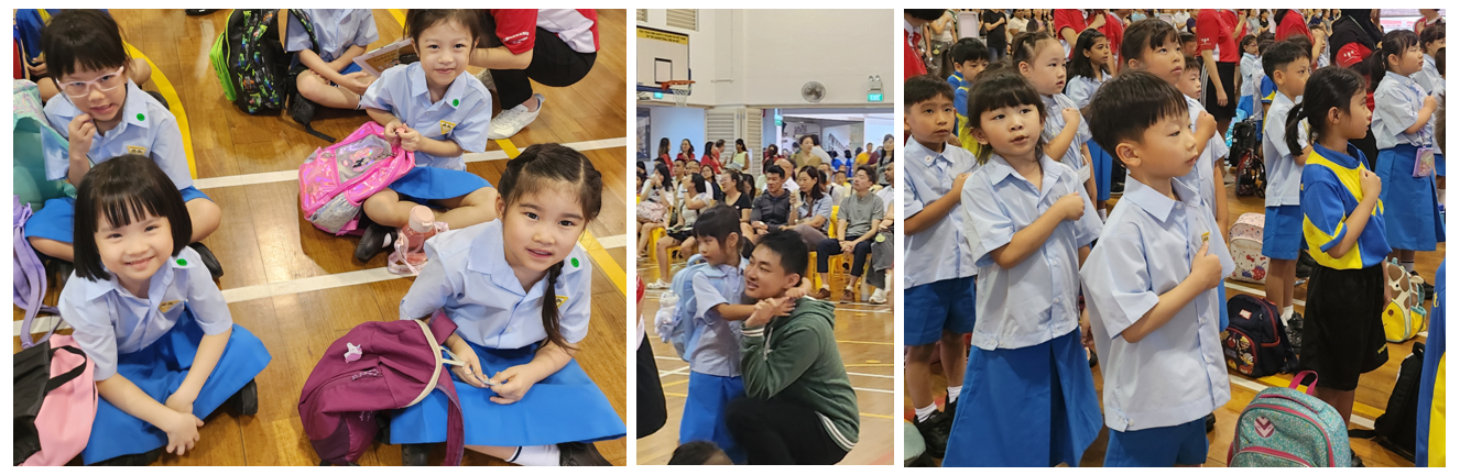 Young children in light blue school uniforms sit and stand in a gymnasium with backpacks during a school assembly.