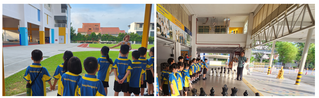 Students in blue and yellow uniforms stand outdoors listening to adults, with giant chess pieces visible on the right.