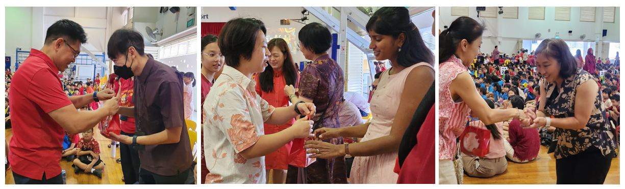 Adults exchange small items at an indoor school event with children seated in the background.
