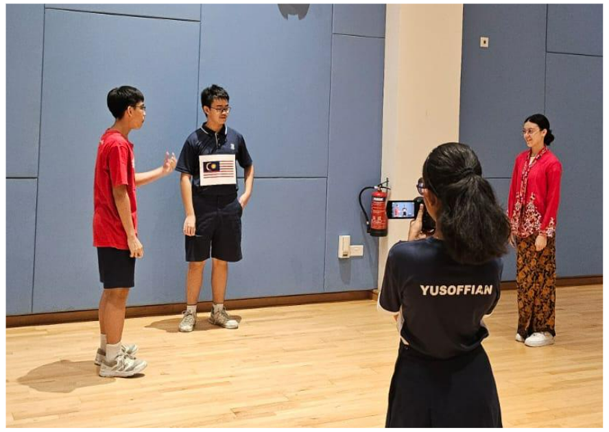 A student films three people in a room, one holding a Malaysian flag, with "YUSOFFIAN" on her shirt.