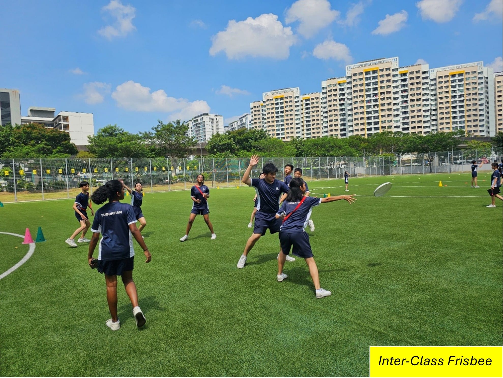 Students in navy uniforms play ultimate frisbee on a green field with apartment buildings in the background.
