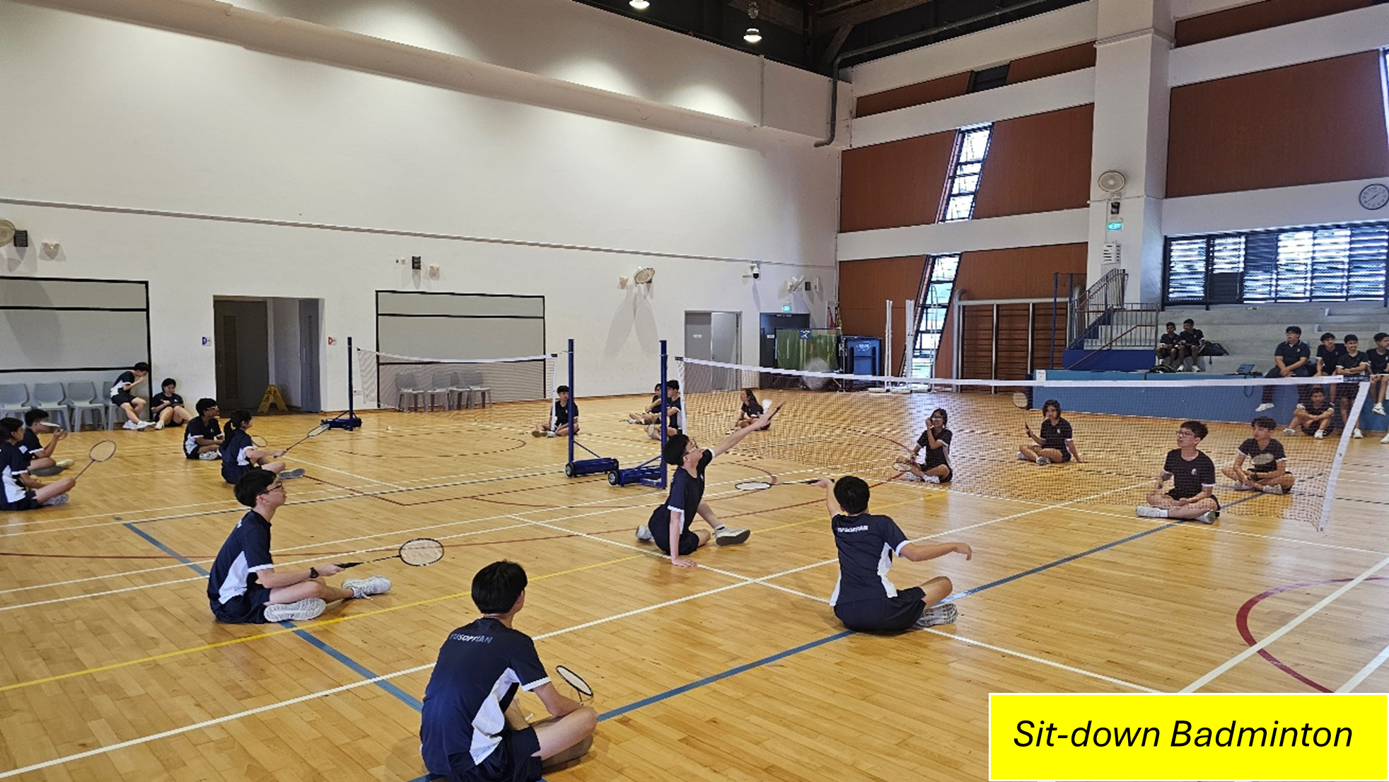 Students sitting on a gym floor play badminton over nets; yellow caption reads Sit-down Badminton.