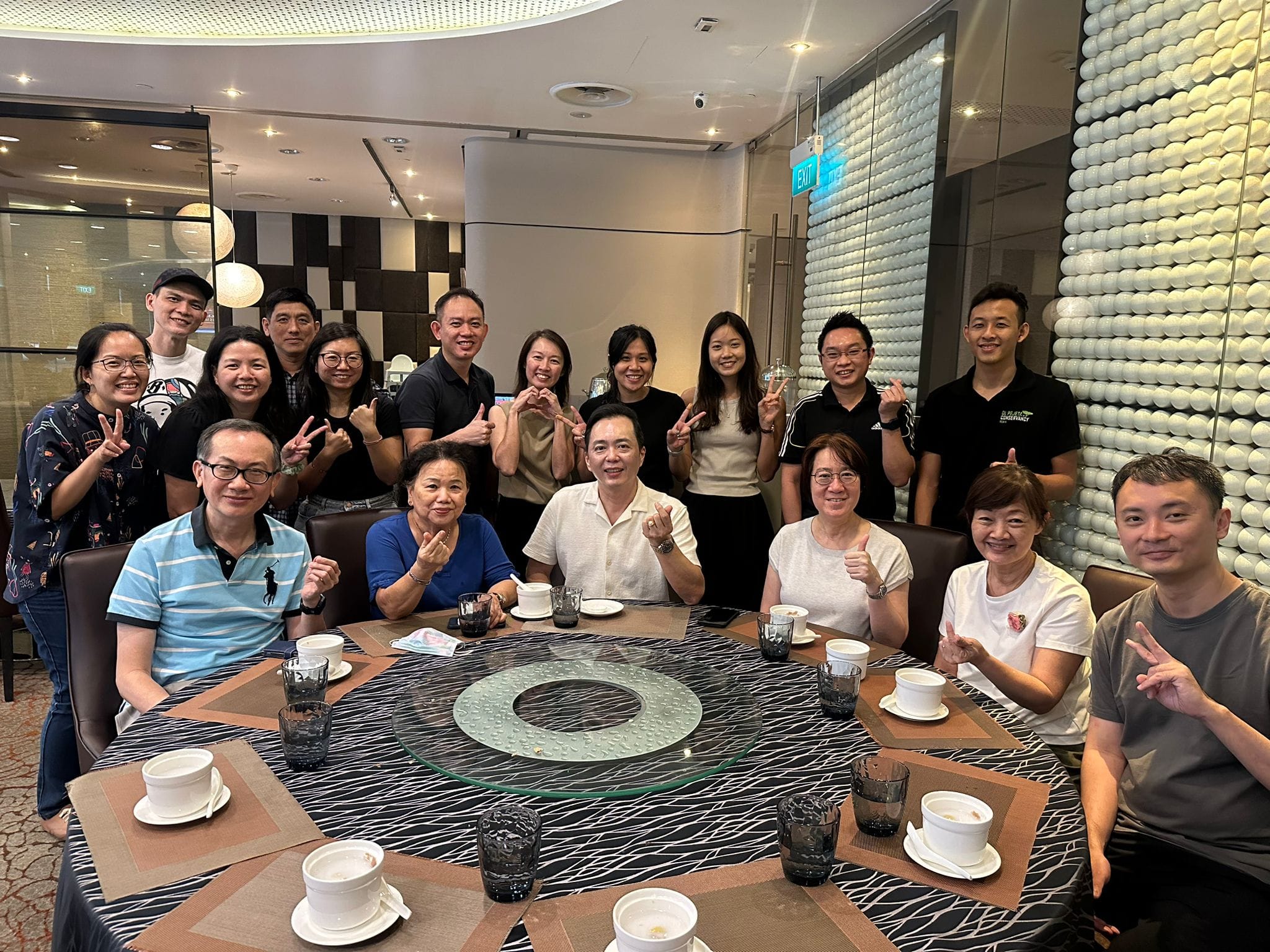 A large group of smiling people posing around a round restaurant table, many making peace or heart signs with their hands.