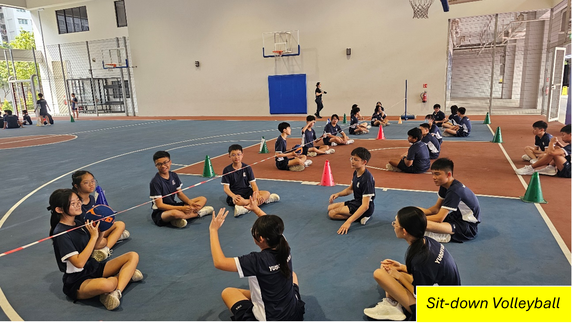 Students in sports uniforms sit on an indoor court, tossing a ball over a low net between cones.